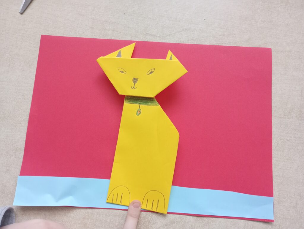 Obrazek przedstawia żółtego kota z papieru na czerwonym tle.