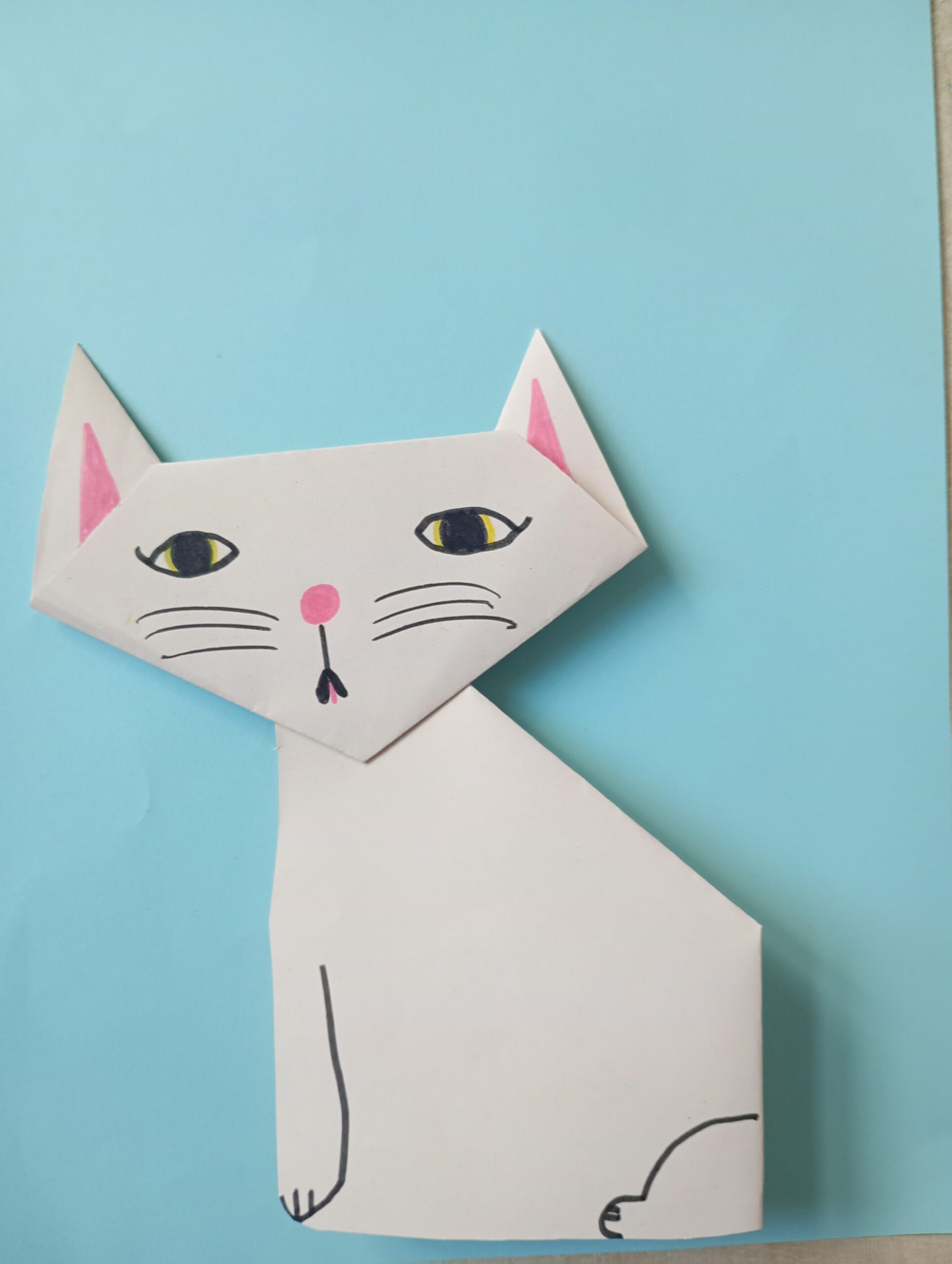 Obrazek przedstawia białego kota wykonanego z papieru na niebieskim tle.